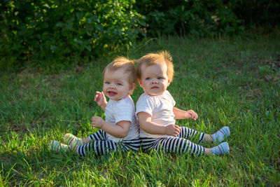 Two twins sit with their backs leaning on the green grass in a city park