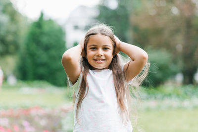 Little girl holds her hands on her head on a walk in the park in summer and smiles