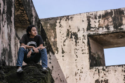 Young man sitting on wall against sky