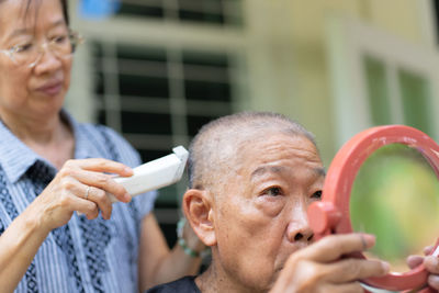 Senior asian wife using trimmer to cut hair of her husband at home patio