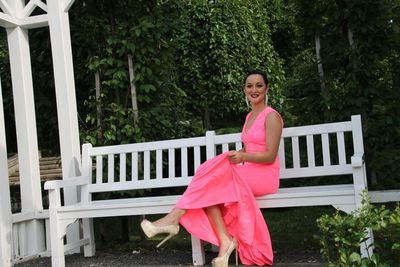 Full length portrait of beautiful bride in wedding dress sitting on white bench in garden