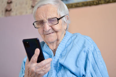 Happy elderly 90-year-old woman with glasses wearing a blue jacket smiles using a smartphone.