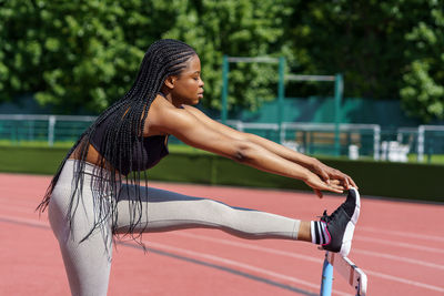 Black young woman with long braids stretches legs and body using hurdles barrier on sports ground