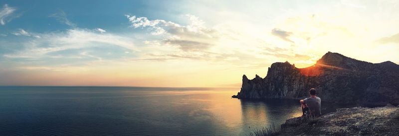 Rear view of man sitting on cliff against sea during sunset