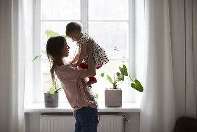 Side view of fashion designer carrying daughter while standing by window at home