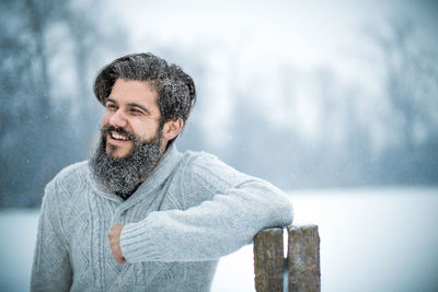 Happy young bearded man with arms crossed standing during snowfall