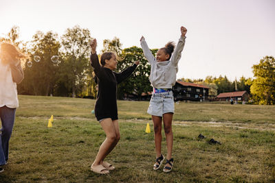 Cheerful girls jumping with arms raised on grass while playing in playground