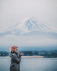 Side view of young woman having drink while standing by lake during winter