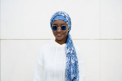 Self assured young african american female millennial in trendy sunglasses and traditional headscarf smiling and looking at camera against white wall