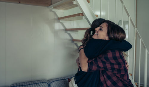 Side view of friends embracing while standing against wall at home