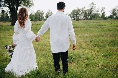 Rear view of bride and groom walking while holding hands grassy field 