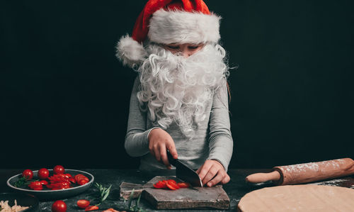 Little girl in a santa hat with a beard cuts with a big knife cherry tomatoes on a gray wooden board