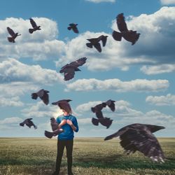 Obscured face of man looking at crows while standing on meadow against sky