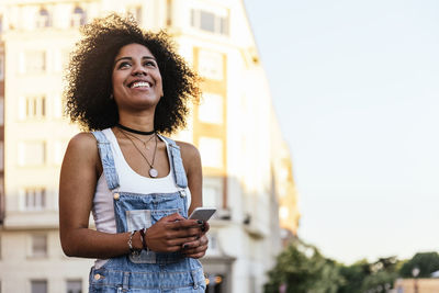 Smiling woman with phone standing against building