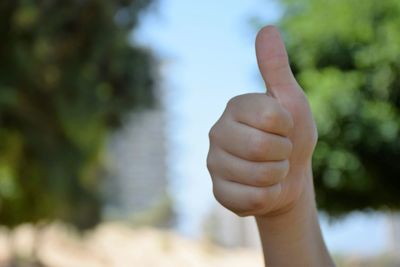 Cropped hand of woman gesturing thumbs up sign against trees