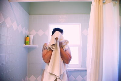 Woman wiping face with towel against window in bathroom