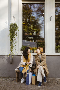 Happy female friends talking to each other while sitting on bench outside store