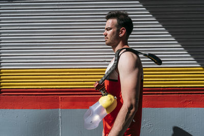 Side view of serious male athlete carrying artificial leg on shoulder standing against painted wall during workout on city street