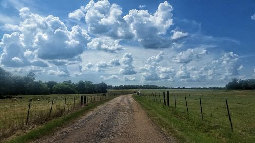 Dirt road passing through field against cloudy sky