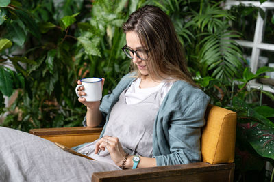 Woman holding coffee cup using smart phone sitting at home