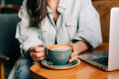 Unrecognizable cropped woman drinking coffee while working on a laptop in a cafe.