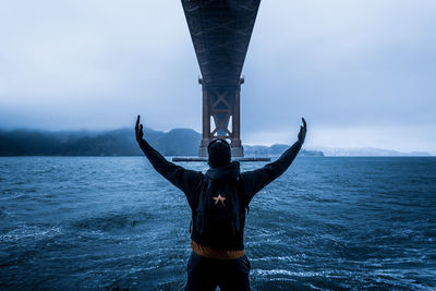 Rear view of man standing under bridge by bay against sky