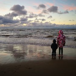Rear view of siblings standing at beach against sky during sunset