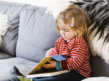 Cute baby girl holding book while sitting on sofa at home