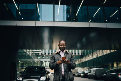 Low angle view of businessman using smart phone while standing on road against building in city