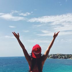 Rear view of woman with arms raised standing by sea against sky