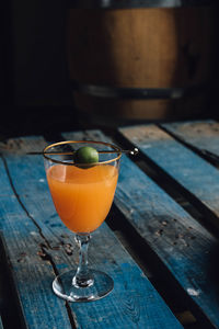 Carrot orange drink in wine glass with green olive on gold cocktail pick on blue wooden pallet