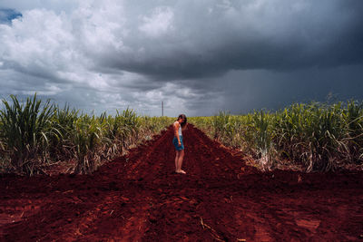 Side view of female traveler in casual wear standing on crossroad with brown soil among green tropical plants under grey cloudy heaven in cuba