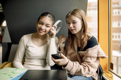 Smiling girl listening to music sitting with female friend holding smart phone at high school