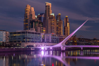 Illuminated buildings by river against sky in city at dusk