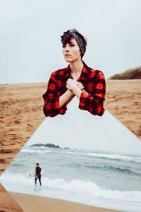 Thoughtful sad young female in casual checkered shirt standing on sandy beach and holding large mirror with reflection of stormy sea and walking man