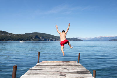 Rear view of woman jumping on pier over lake against sky