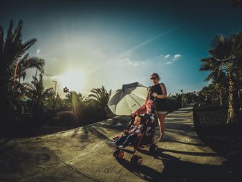 Mother with baby in carriage on footpath against sky during sunny day