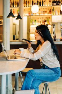 Side view of young ethnic female sending voice message on cellphone at cafeteria table with netbook