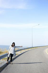 Full length of woman hitchhiking while standing on road against sky