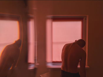 Rear view of shirtless man standing by window at home