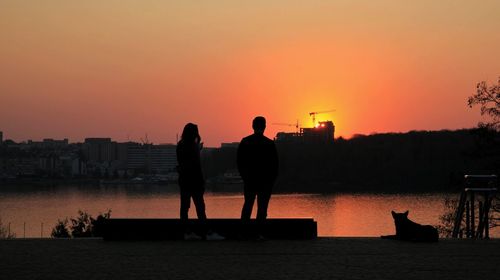 Silhouette people standing on shore against orange sky during sunset