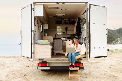 Delighted female traveler sitting outside on stairs of van while working remotely on laptop