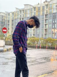 Side view of teenage boy wearing protective mask while standing on wet street in city