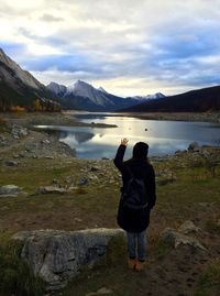 Rear view of woman with hand raised standing on field by lake against mountains