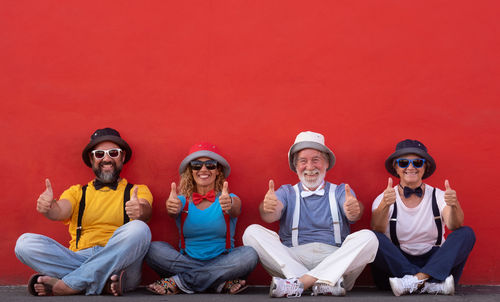 Portrait of happy friends showing thumbs up while sitting against red wall