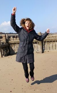 Portrait of happy senior woman jumping on beach against sky