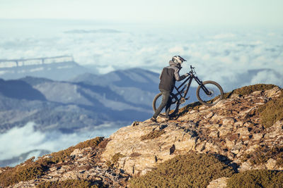 Side view of guy with helmet upping bicycle to peak of hill and picturesque view of mountains in clouds