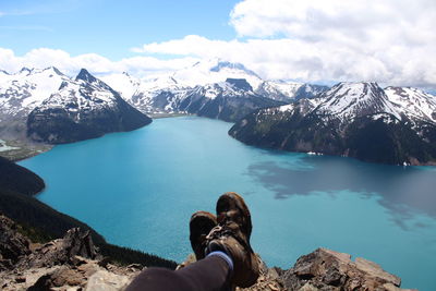 Low section of man sitting on cliff by lake and snowcapped mountains against sky