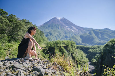 Full length of woman looking away while crouching on rock against mountains and sky