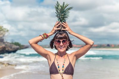 Portrait of seductive women holding pineapple at beach against sky